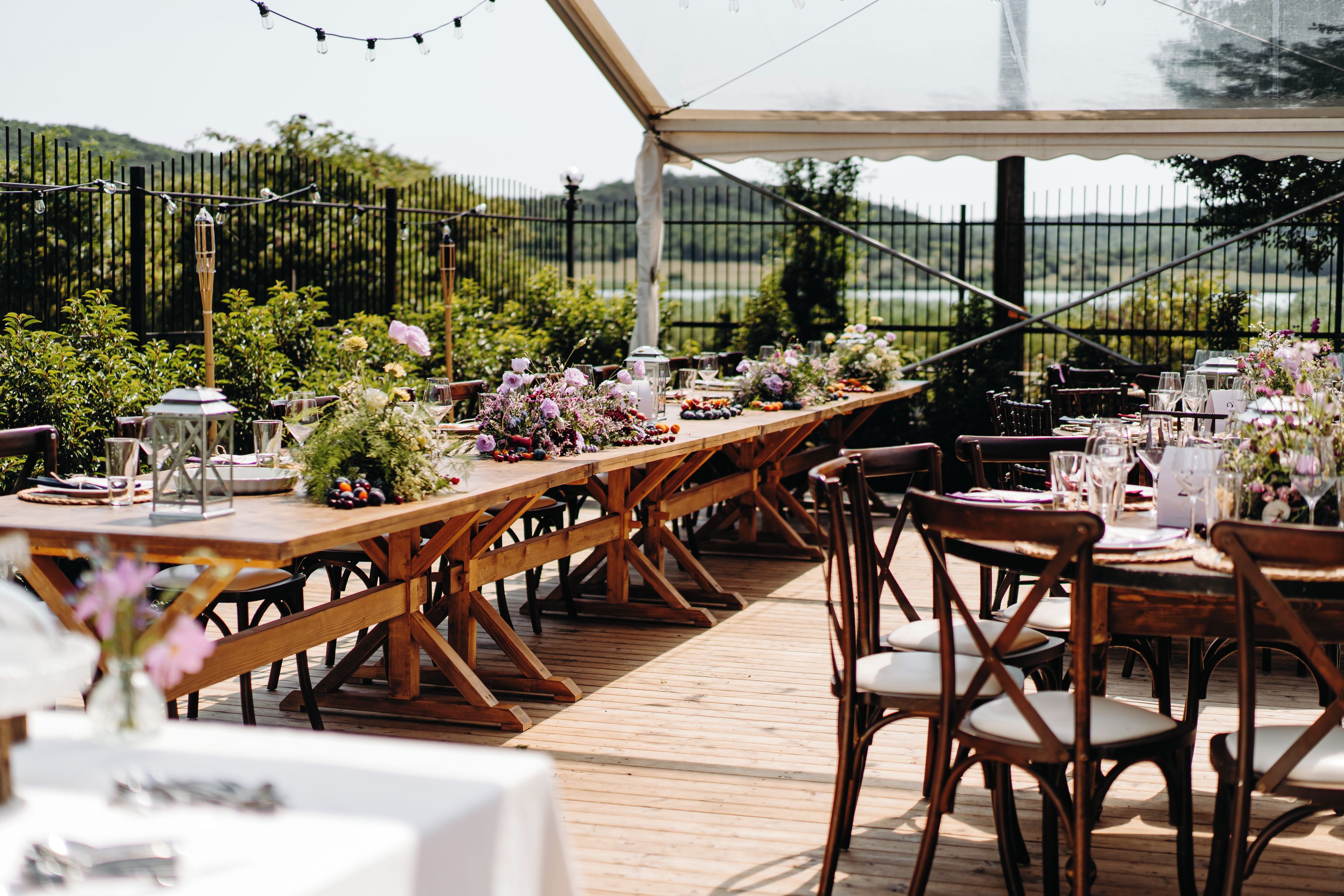 An outdoor dining area with wooden tables and chairs, surrounded by greenery and a view of the countryside.