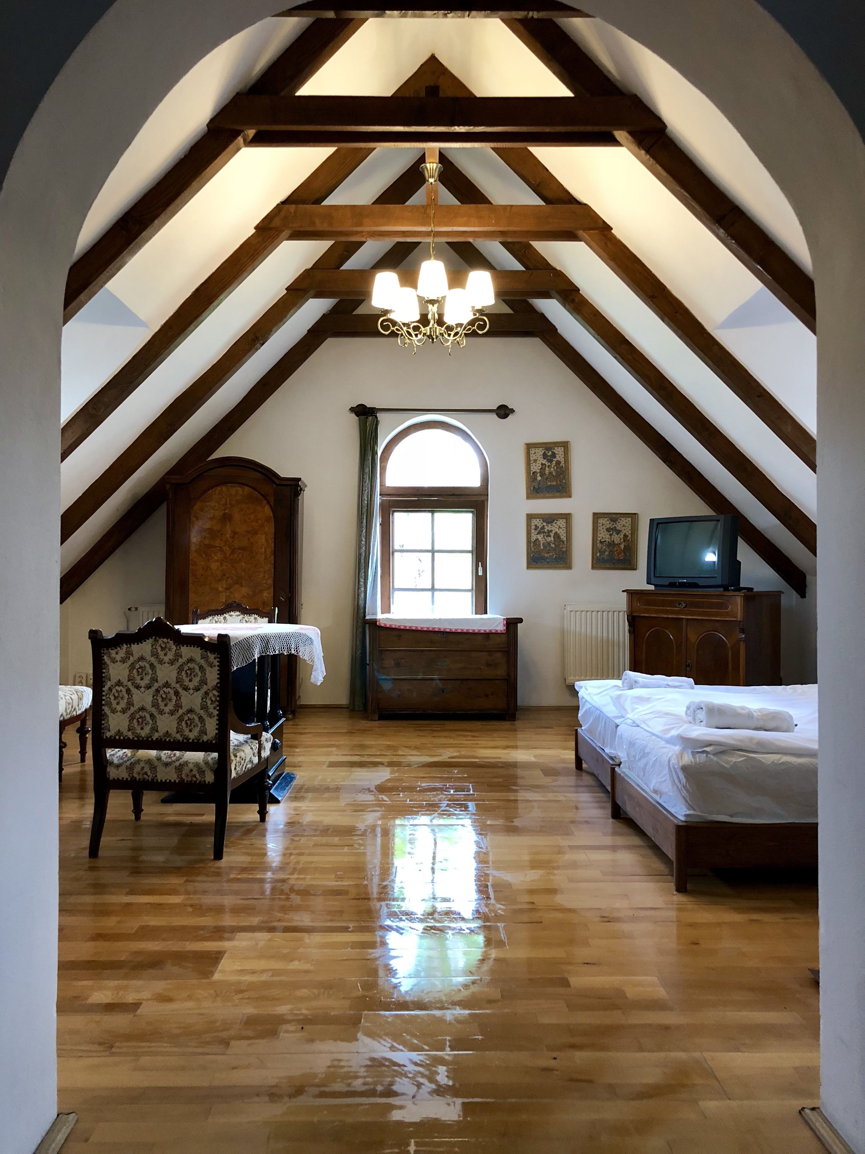 A room with a sloped ceiling, wooden beams, a chandelier, a bed, a chair, a table, and a television.