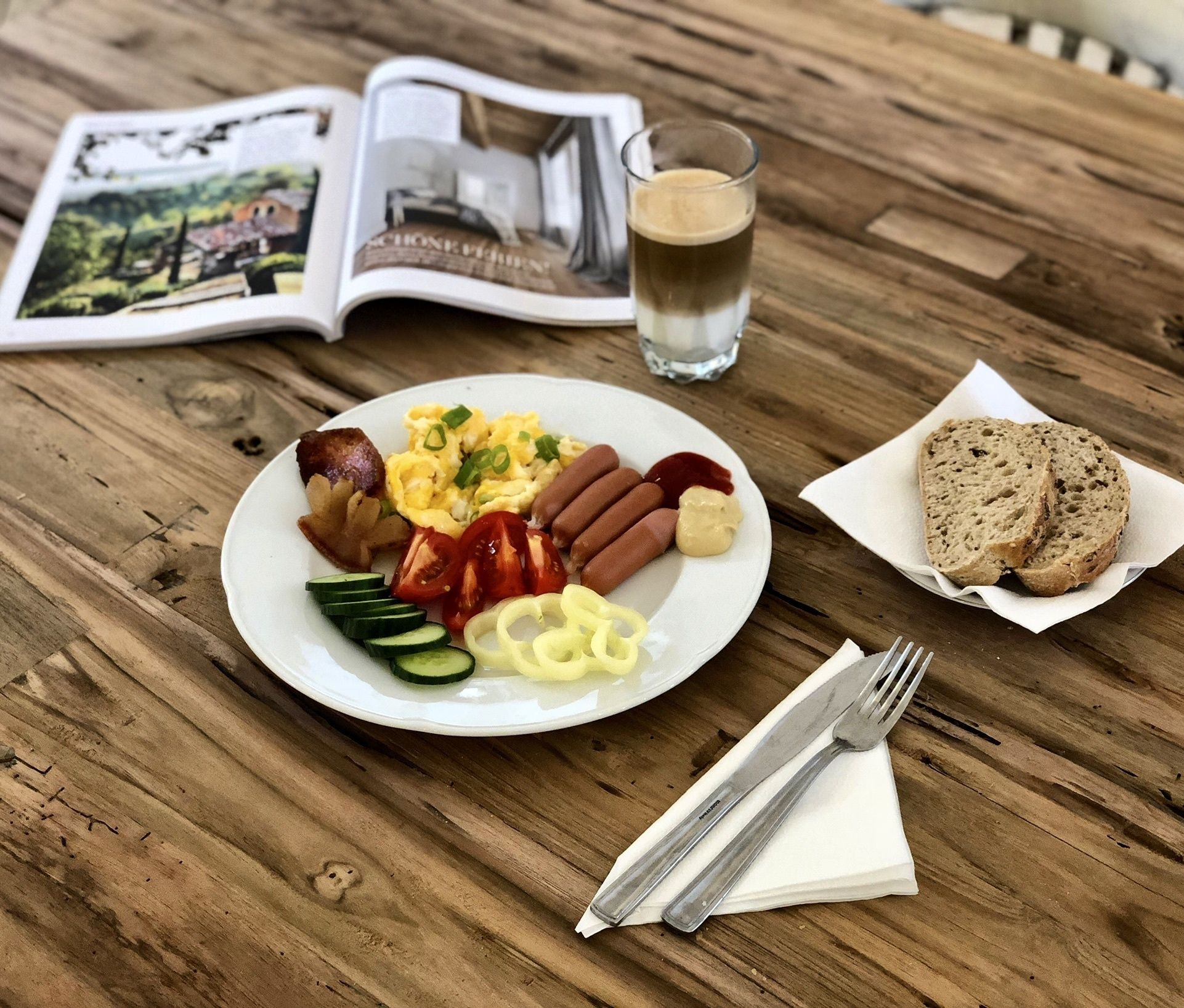 Un plato con salchichas, huevos y verduras, una taza de café y un libro sobre una mesa de madera.