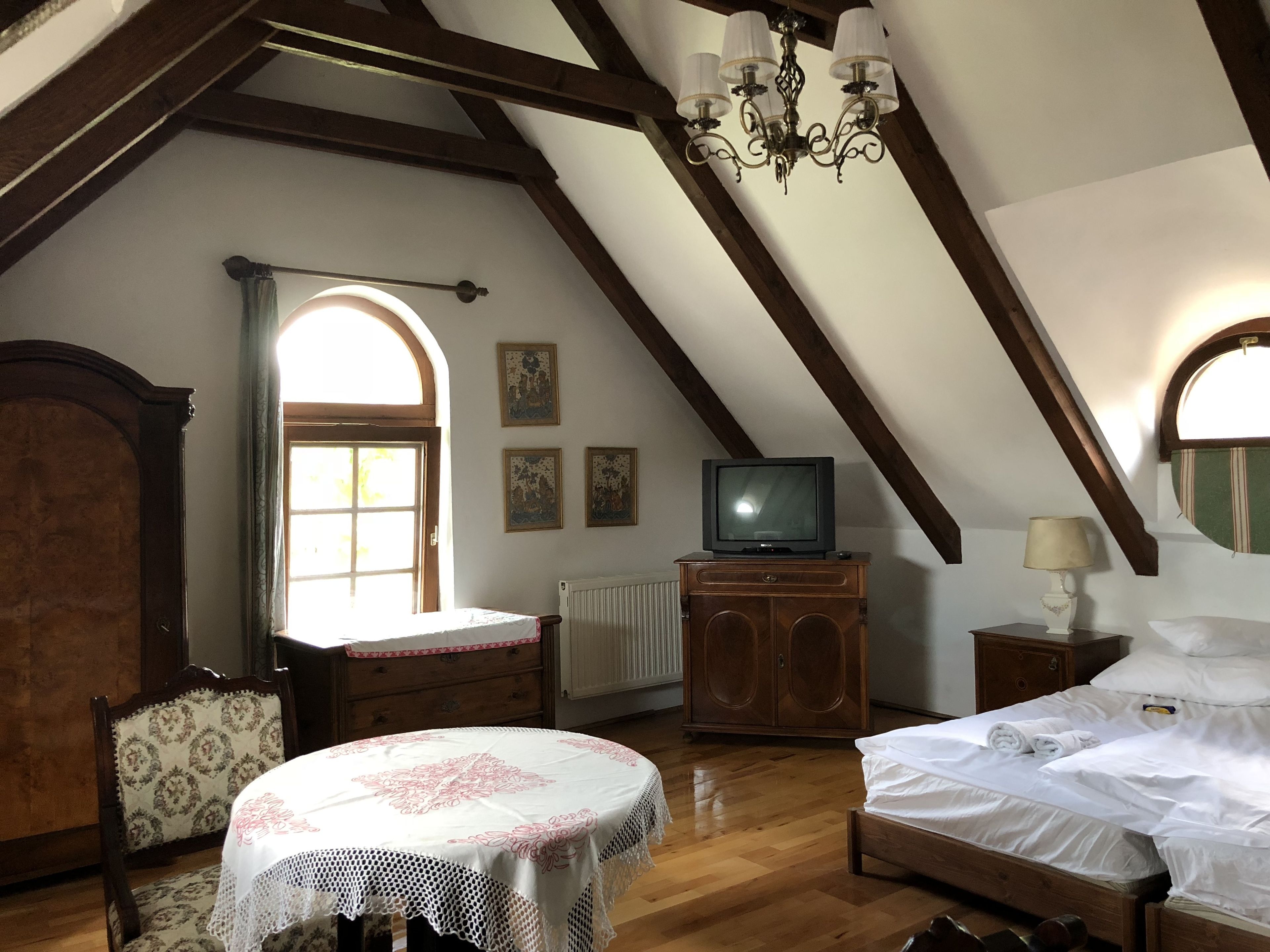 A bedroom with a wooden bed, a small table with a lace tablecloth, a television on a cabinet, and a window with curtains.