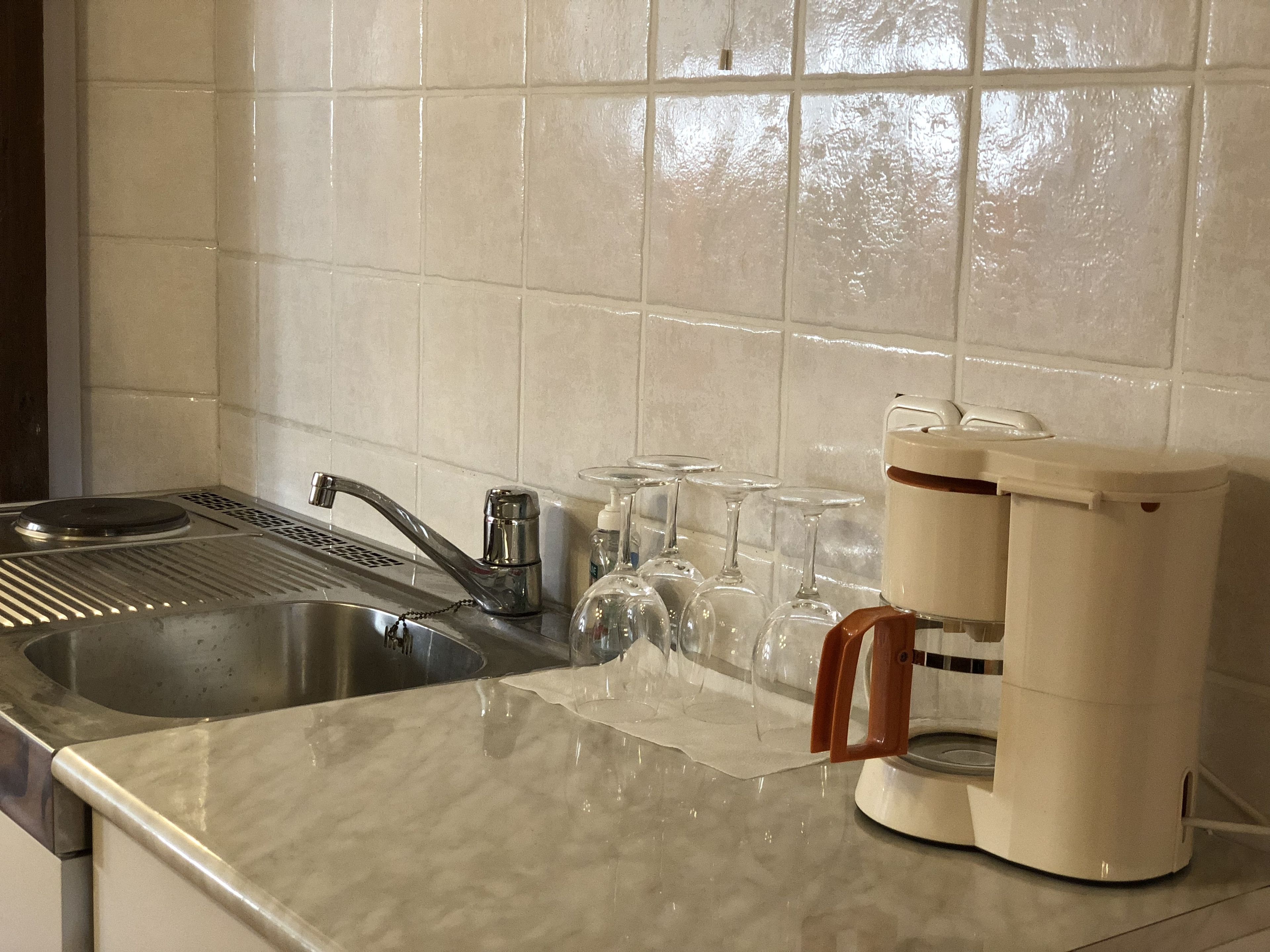 Una cocina con un lavamanos de acero inoxidable, una cafetera y varias copas de vino.