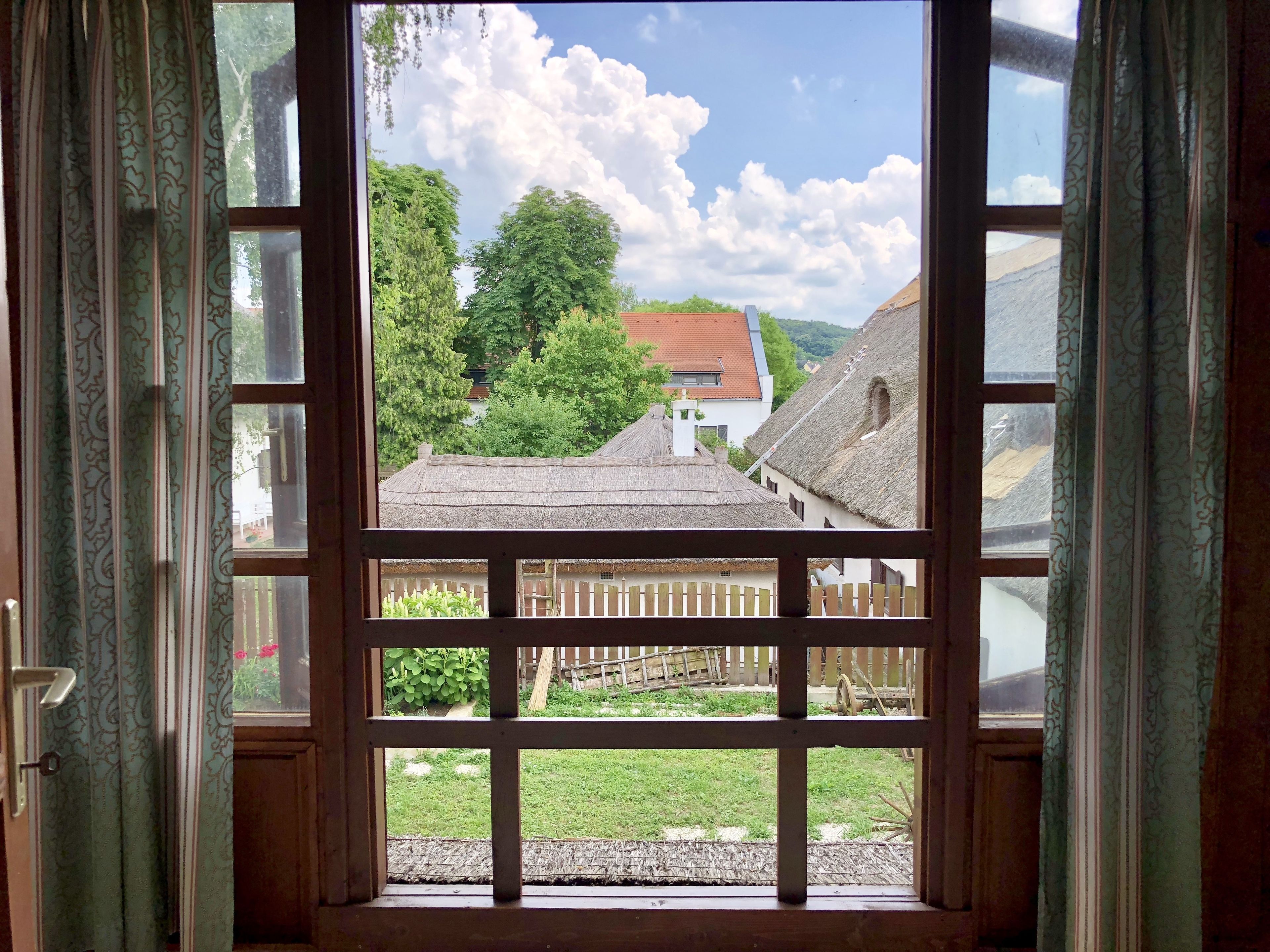 A view through a window with a wooden frame, revealing a rural landscape with thatched-roof buildings, a wooden fence, and greenery.