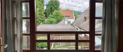 A view through a window with a wooden frame, revealing a rural landscape with thatched-roof buildings, a wooden fence, and greenery.