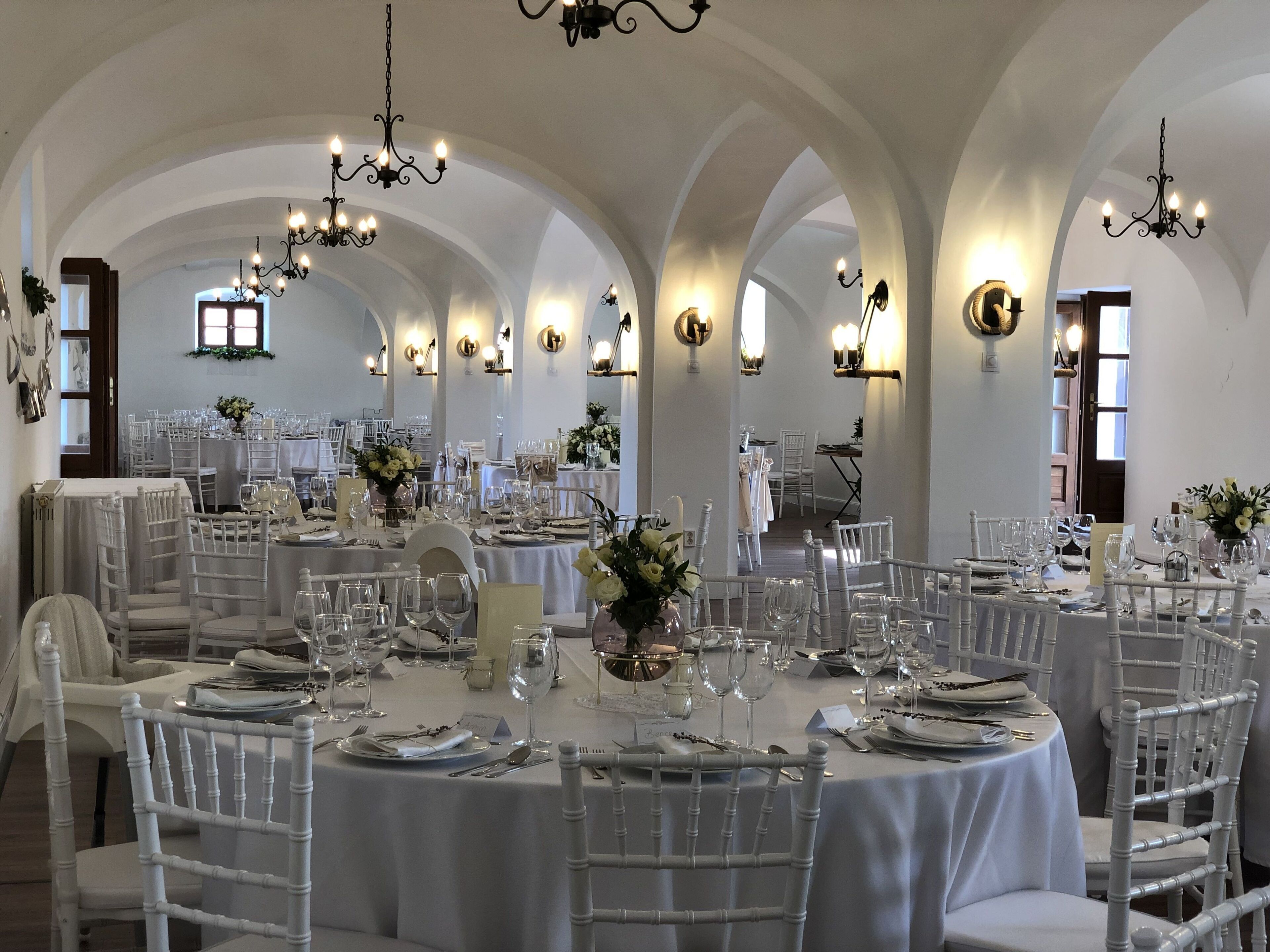 A banquet hall with round tables, white chairs, chandeliers, and wall sconces.