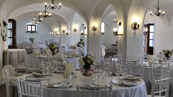A banquet hall with round tables, white chairs, chandeliers, and wall sconces.