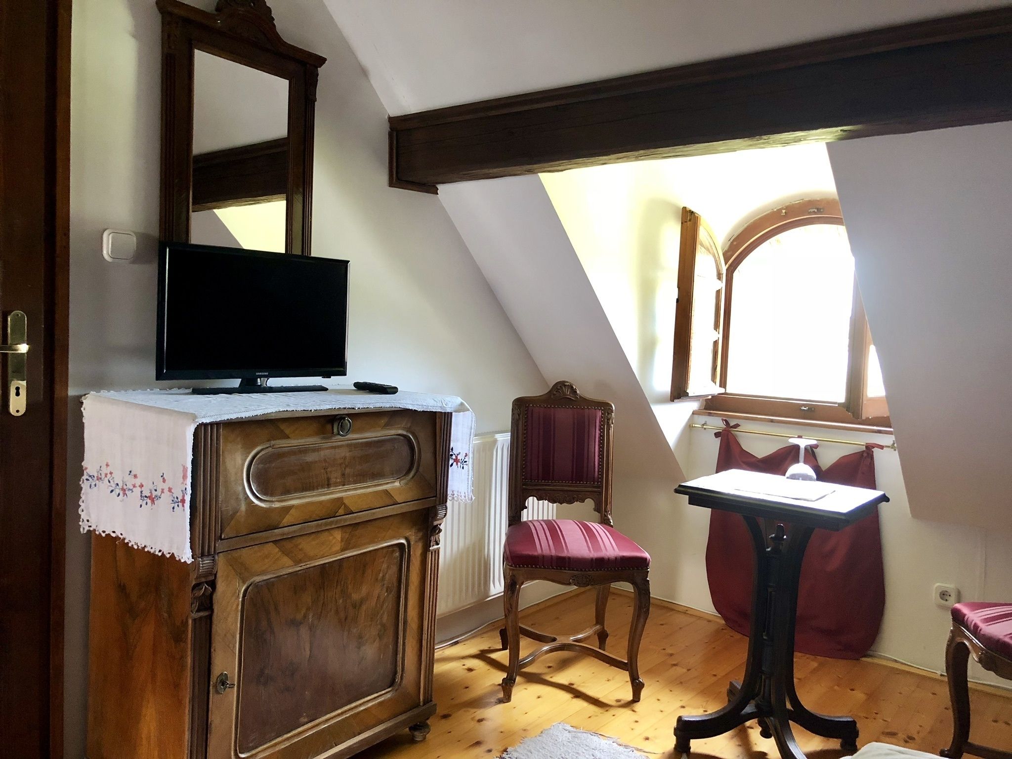 A room with a wooden dresser, a chair, a table, a television, and a window with curtains.