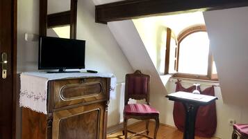 Ein Zimmer mit einem Holzkoffer, einem Sessel, einem Tisch, einem Fernseher und einem Fenster mit Vorhängen.