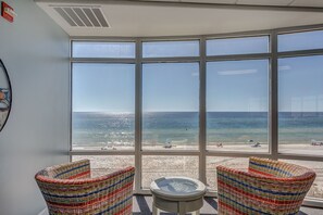 A room with large windows overlooking a beach, two wicker chairs, a small round table, and a fire alarm on the wall. - 2 Bedroom in Emerald Isle Resort -  Newly Remodeled Amazing Views (Panama City Beach)