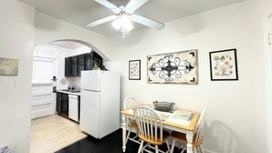 Une cuisine de taille modeste, avec des meubles blancs, un réfrigérateur blanc et une table à manger accompagnée de deux chaises. Un ventilateur de plafond est installé au-dessus de la cuisine, et des œuvres d’art sont accrochées aux murs.