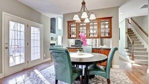 A dining area with a round table, green chairs, and a chandelier. There is a kitchen with wooden cabinets and a staircase in the background.