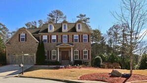 A two-story brick house with a front garage, a central entrance with a door, and a landscaped front yard with red mulch and a tree.