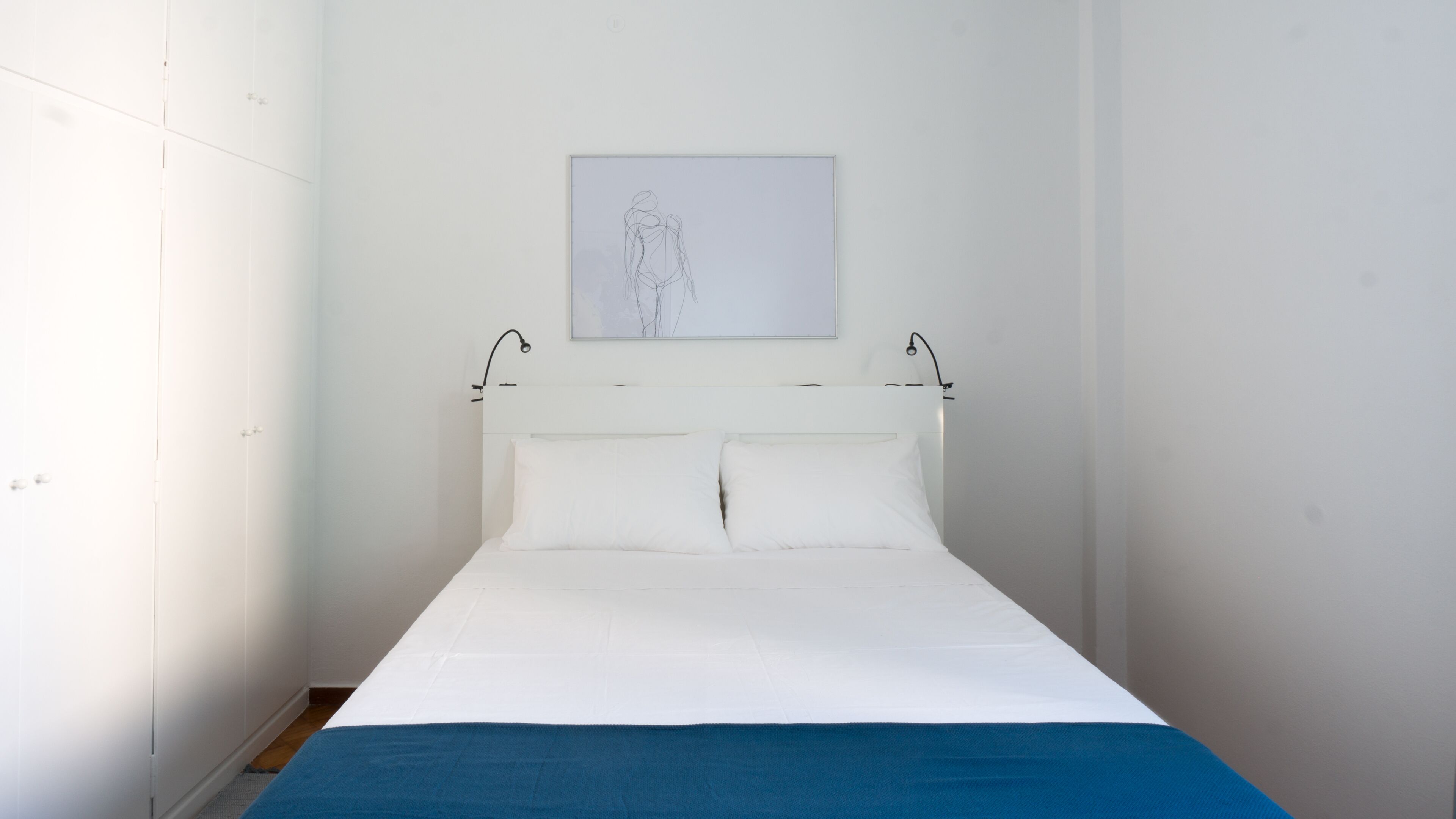 Een kleine, witte kamer met een enkel bed, een blauwe deken, witte kussens en een eenvoudig, modern design.