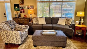 Una sala de estar con un sofá seccional gris, un sillón estampado y una mesa de centro de madera. Hay dos lámparas sobre mesitas auxiliares y una ventana con persianas.