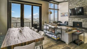 A spacious living area with a large wooden dining table, comfortable seating, a stone fireplace, and a large window offering a scenic view. - Schweitzer Luxury Ski in/out Townhome | Hot Tub (Sandpoint)