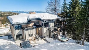 A modern, two-story cabin with a glass facade, a hot tub, and a view of snow-covered mountains. - Schweitzer Luxury Ski in/out Townhome | Hot Tub (Sandpoint)