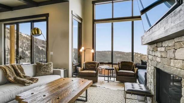 A cozy living room with a stone fireplace, large windows offering mountain views, a wooden coffee table, and leather armchairs. - Schweitzer Luxury Ski in/out Townhome | Hot Tub (Sandpoint)