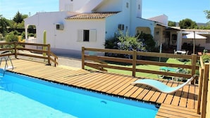 A pool area with a wooden deck, a lounge chair, and a white building with a tiled roof in the background. - Villa in Algarve with complete privacy (moncarapacho,)