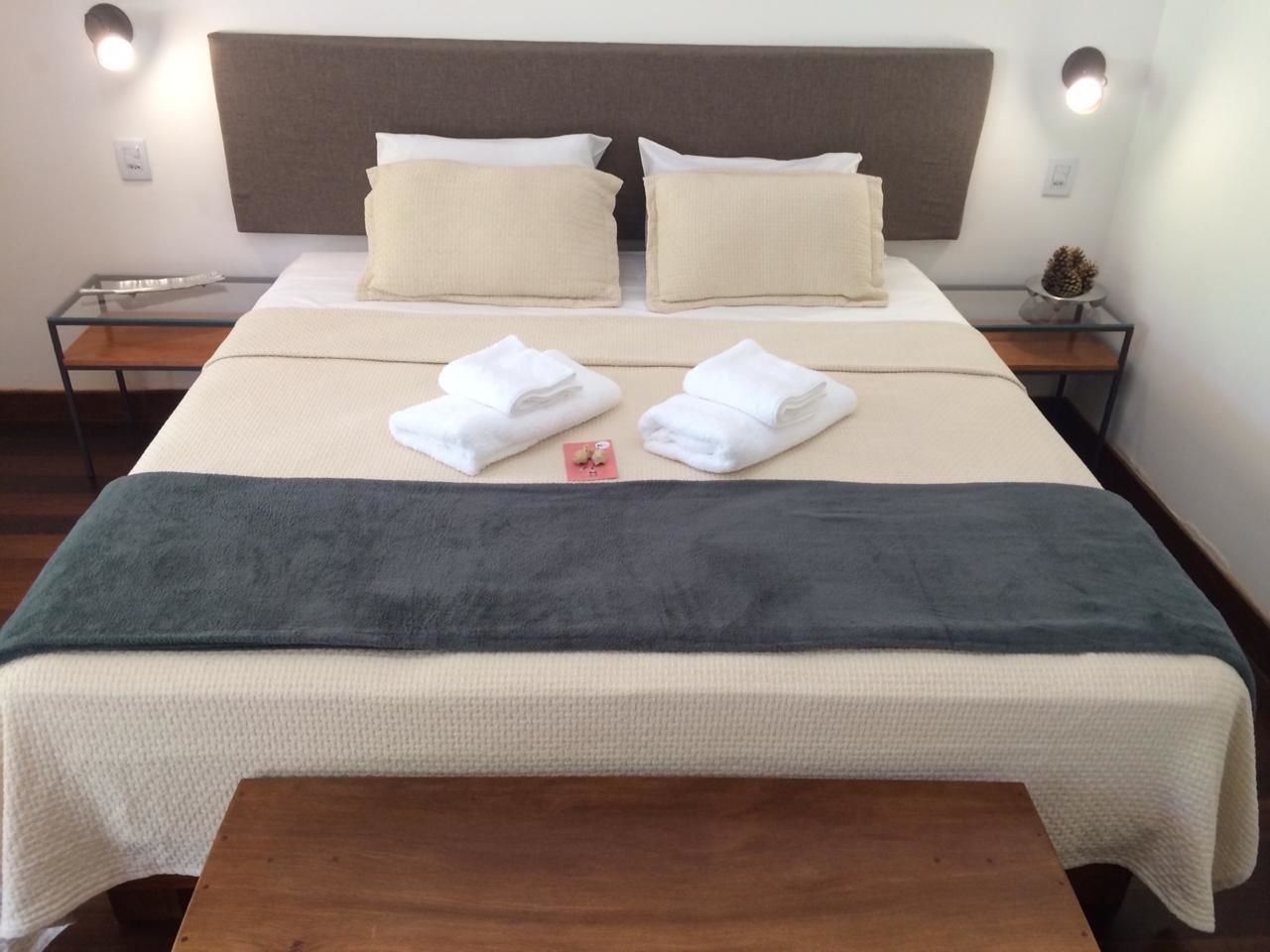 Un lit bien fait, recouvert d’une couverture grise, avec deux oreillers et des serviettes pliées posées sur une table de chevet en bois.