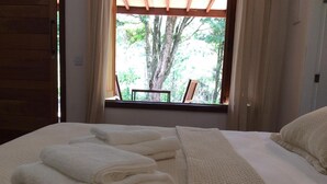 A neatly made bed with folded white towels and a view of trees through a window.