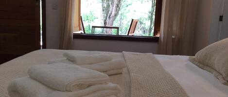 A neatly made bed with folded white towels and a view of trees through a window.