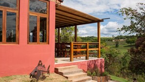 A modern house with a red exterior, large windows, a wooden porch, and a view of a green landscape.