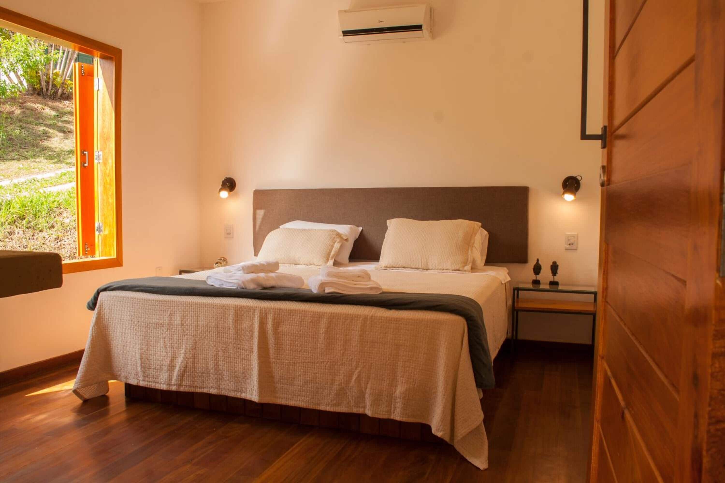 A bedroom with a bed, bedside tables, an air conditioning unit, and a window with a view of greenery.