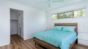 A bedroom with a wooden bed, a nightstand, a ceiling fan, and a window with a view of trees. - 25 Cooloola Drive - Rainbow Beach. Stunning, Modern, Open Plan Beach House. Wifi. (RAINBOW BEACH)