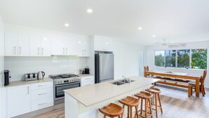 A modern kitchen with white cabinets, stainless steel appliances, a central island with stools, and a dining area with a view through a large window. - 25 Cooloola Drive - Rainbow Beach. Stunning, Modern, Open Plan Beach House. Wifi. (RAINBOW BEACH)