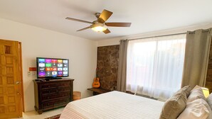 Une chambre à coucher avec un lit, un téléviseur posé sur une commode, une guitare et un ventilateur de plafond.