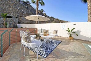 An outdoor patio with white metal chairs and a table, a beige umbrella, and a patterned rug.