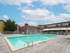A rectangular pool with a wooden deck, surrounded by lounge chairs and umbrellas, with buildings in the background. - Appart sympa proche du Golf, accès Piscine (Rivières)