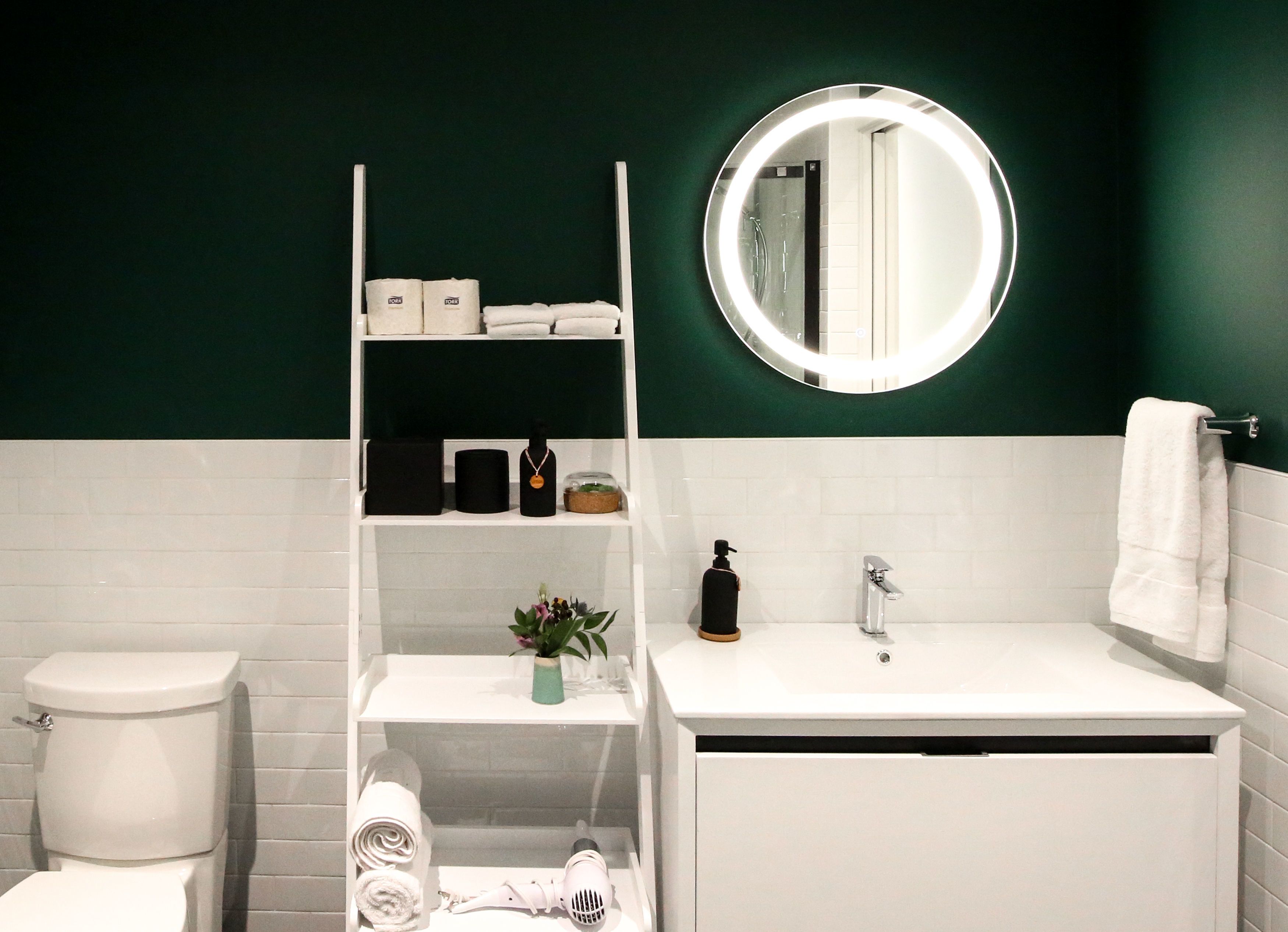 Un baño moderno con un lavabo blanco, un espejo redondo, un inodoro y un estante con artículos de aseo.