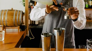 A bartender preparing a drink with a shaker and a bottle, with a wooden bar counter and a wine rack in the background. - Pluvio Restaurant and Rooms (Ucluelet)
