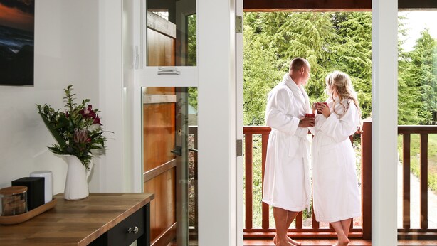 A couple in white robes on a wooden deck, a wooden table with a vase of flowers, and a view of greenery. - Pluvio Restaurant and Rooms (Ucluelet)