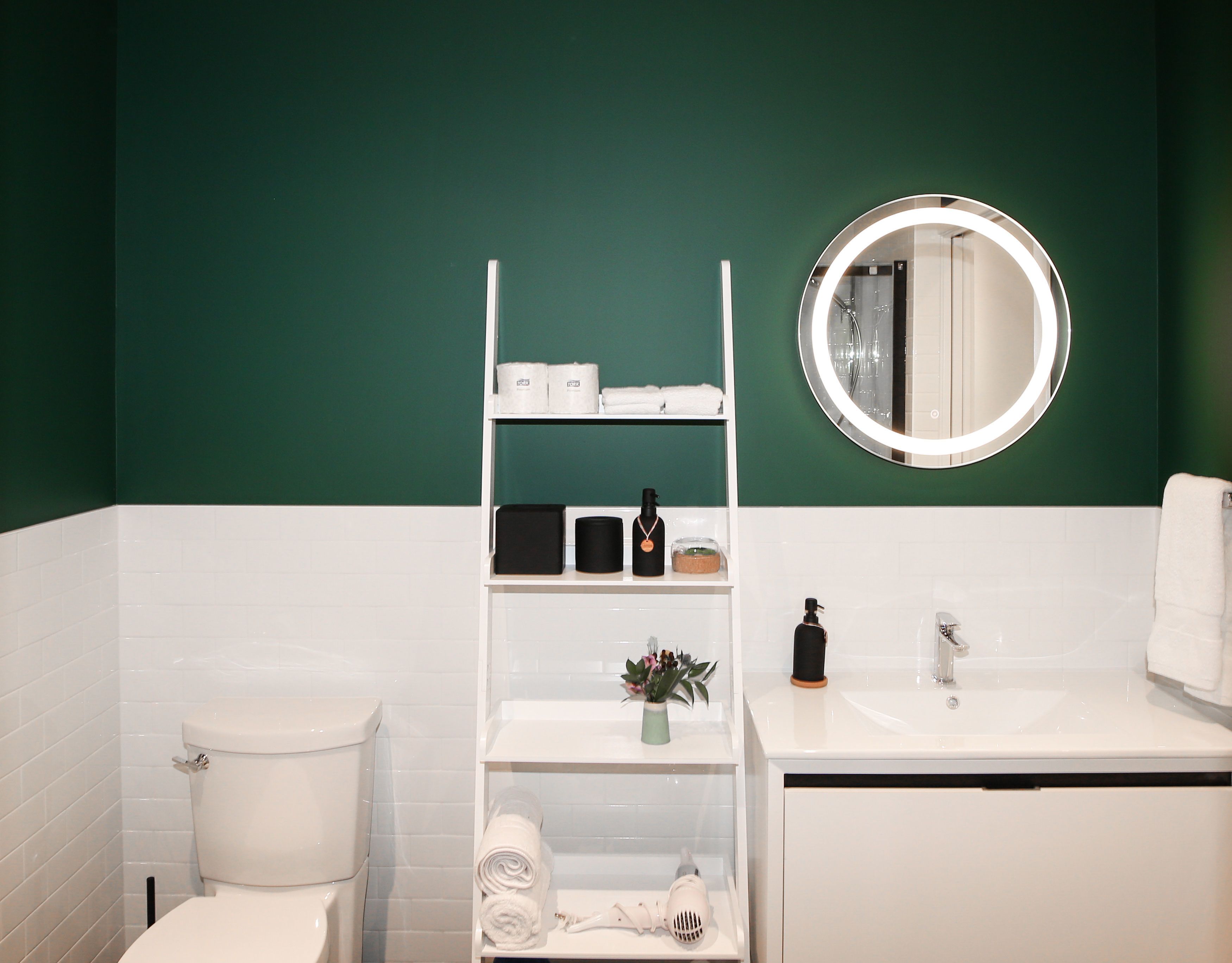 Un baño con inodoro blanco, lavamanos, espejo y estantes con artículos de aseo.