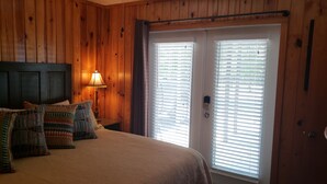 Een slaapkamer met een bed, kussens, een nachtkastje, een lamp en een deur met luiken.
