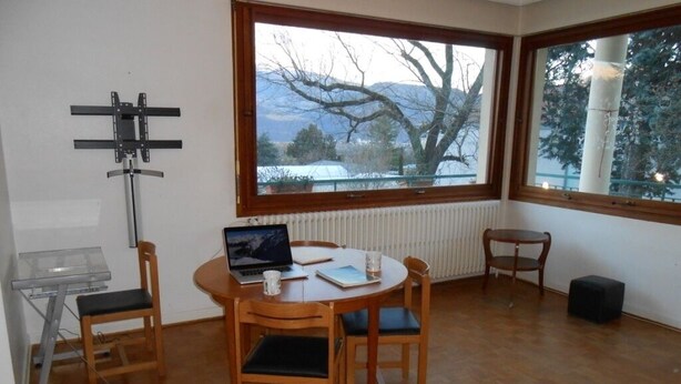 Una habitación con una mesa redonda de madera, cuatro sillas, una computadora portátil, un vaso y un cuaderno. Hay un televisor sobre un mueble, un radiador y una ventana que da a vistas de árboles y montañas.