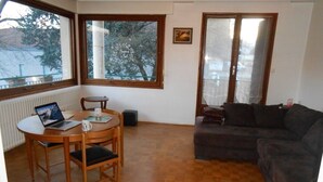 Una sala de estar con piso de madera, una mesa redonda para comer con sillas, un sofá de color oscuro y una ventana con vistas a árboles y edificios.