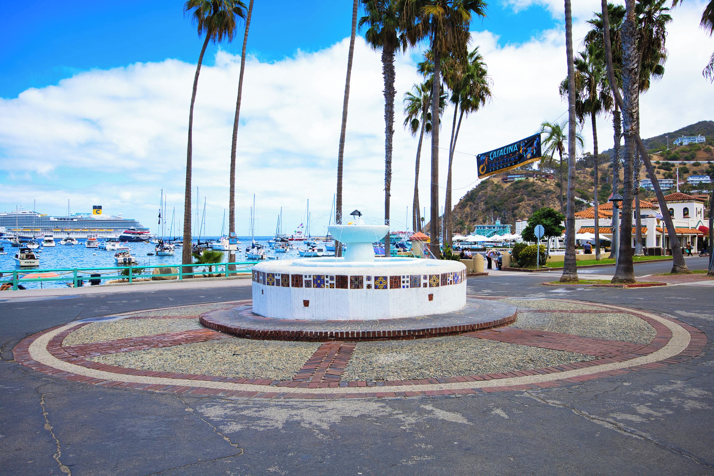 Una plaza frente al mar con una fuente circular, palmeras y un crucero amarrado al fondo.