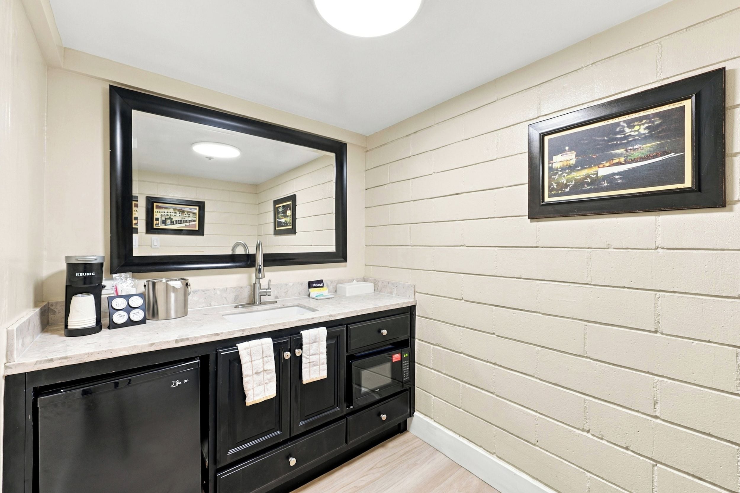 A modern bathroom with a large mirror, black cabinets, a coffee maker, and a framed picture on the wall.