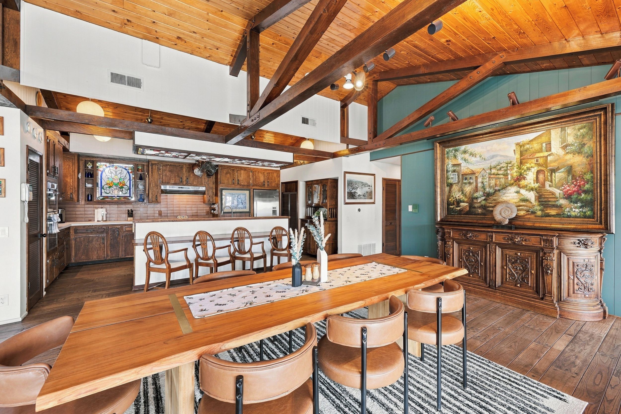 A spacious dining area with a wooden table and chairs, a kitchen in the background, and a large painting on the wall.