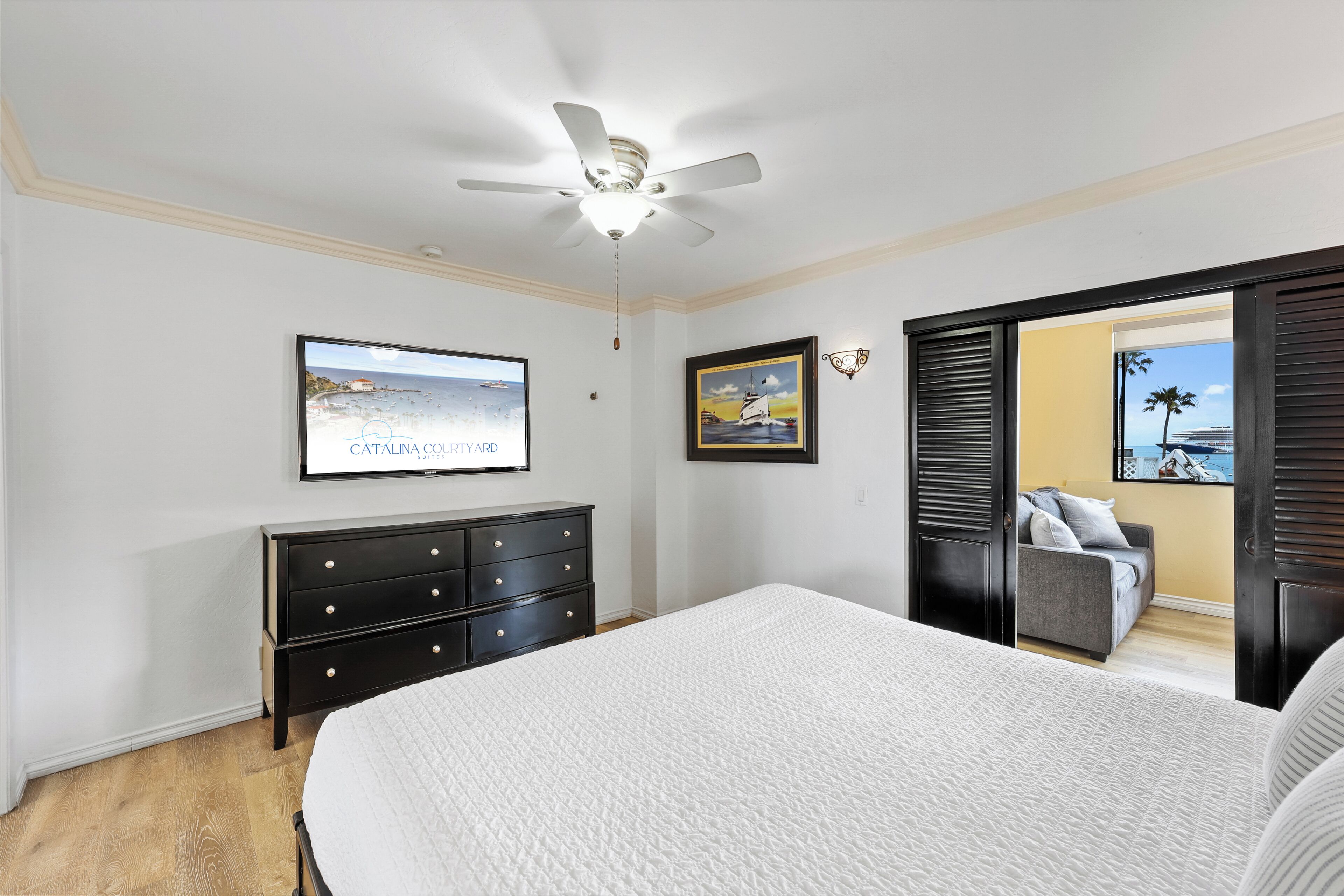 A bedroom with a bed, a ceiling fan, a TV, a dresser, and a view of a balcony with a chair.