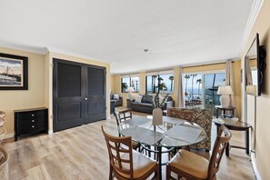 A spacious living area with a glass dining table, comfortable seating, and large windows offering a view of the ocean. - Catalina Courtyard Suites (Avalon)