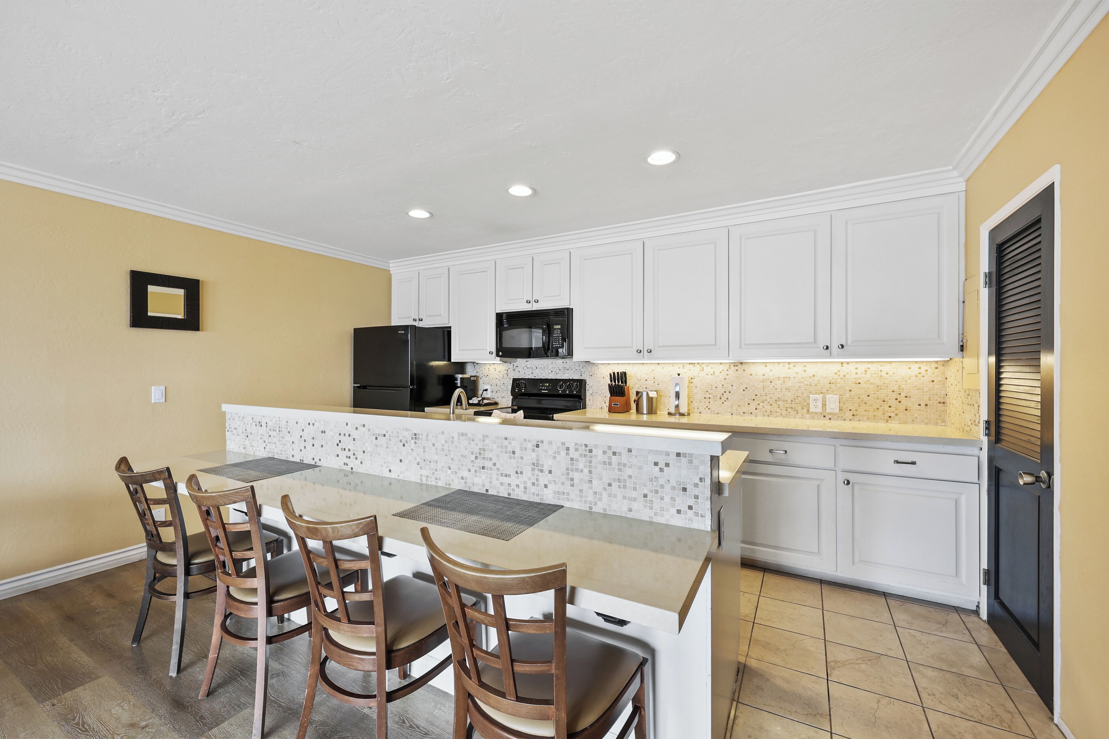 A modern kitchen with white cabinets, a central island with chairs, a microwave, and a refrigerator.
