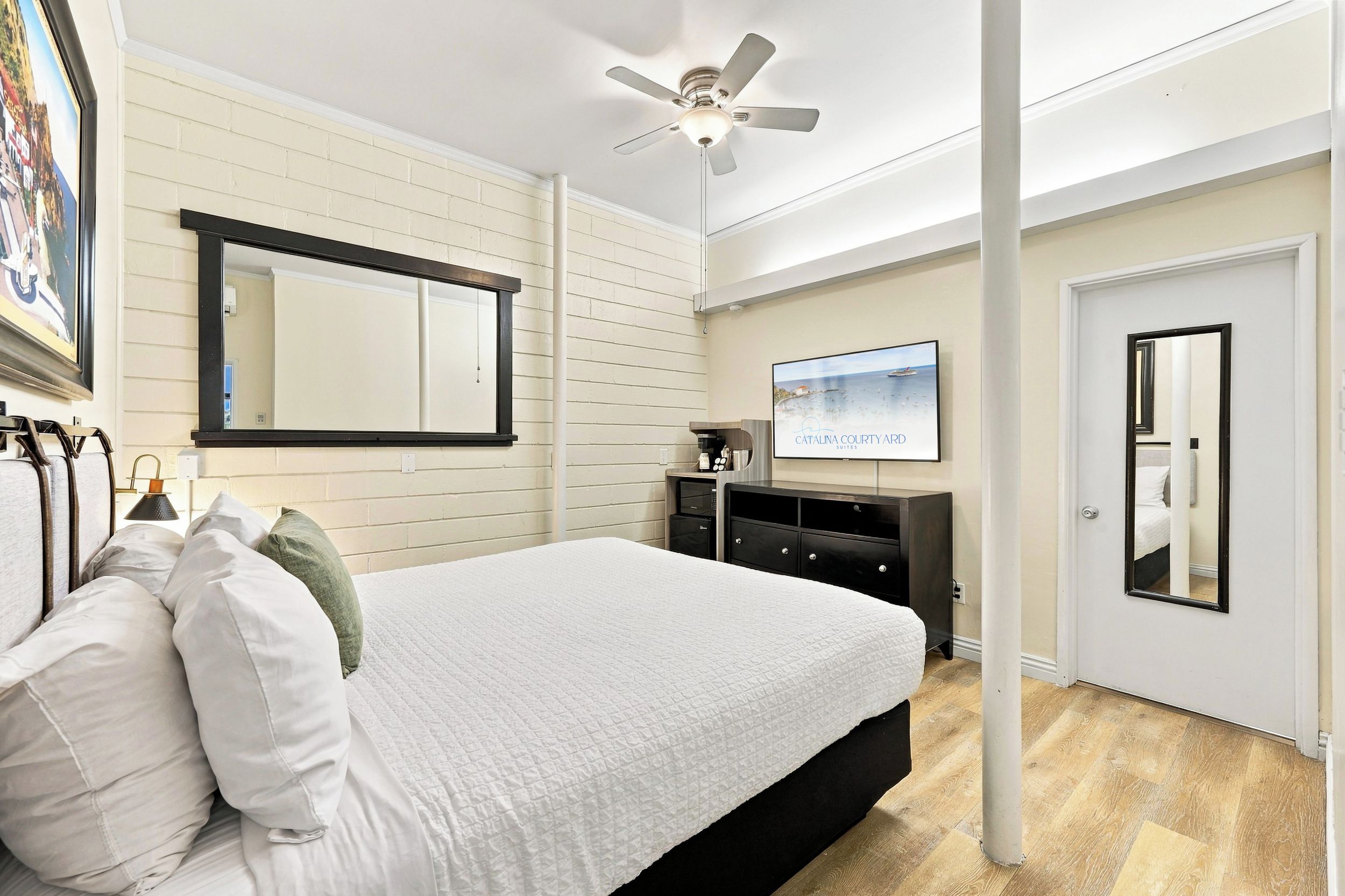 Habitación de hotel con cama, ventilador de techo, televisor de pantalla plana, un espejo grande y una puerta.