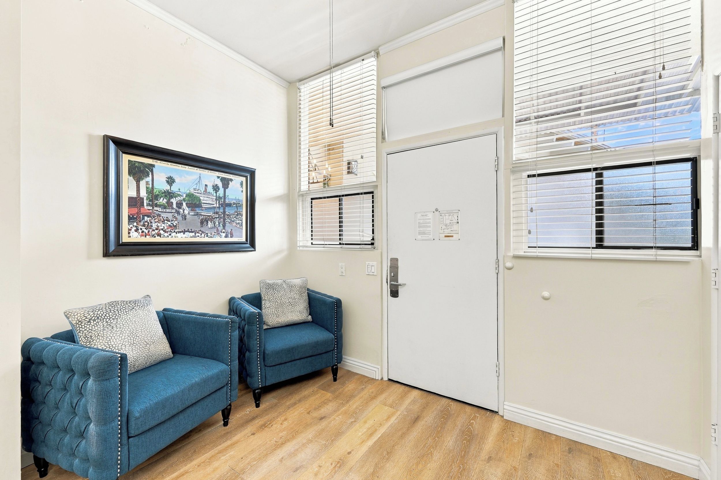 Une pièce avec deux fauteuils bleus, un tableau encadré au mur et une porte blanche sur laquelle est fixé un tableau d’affichage.