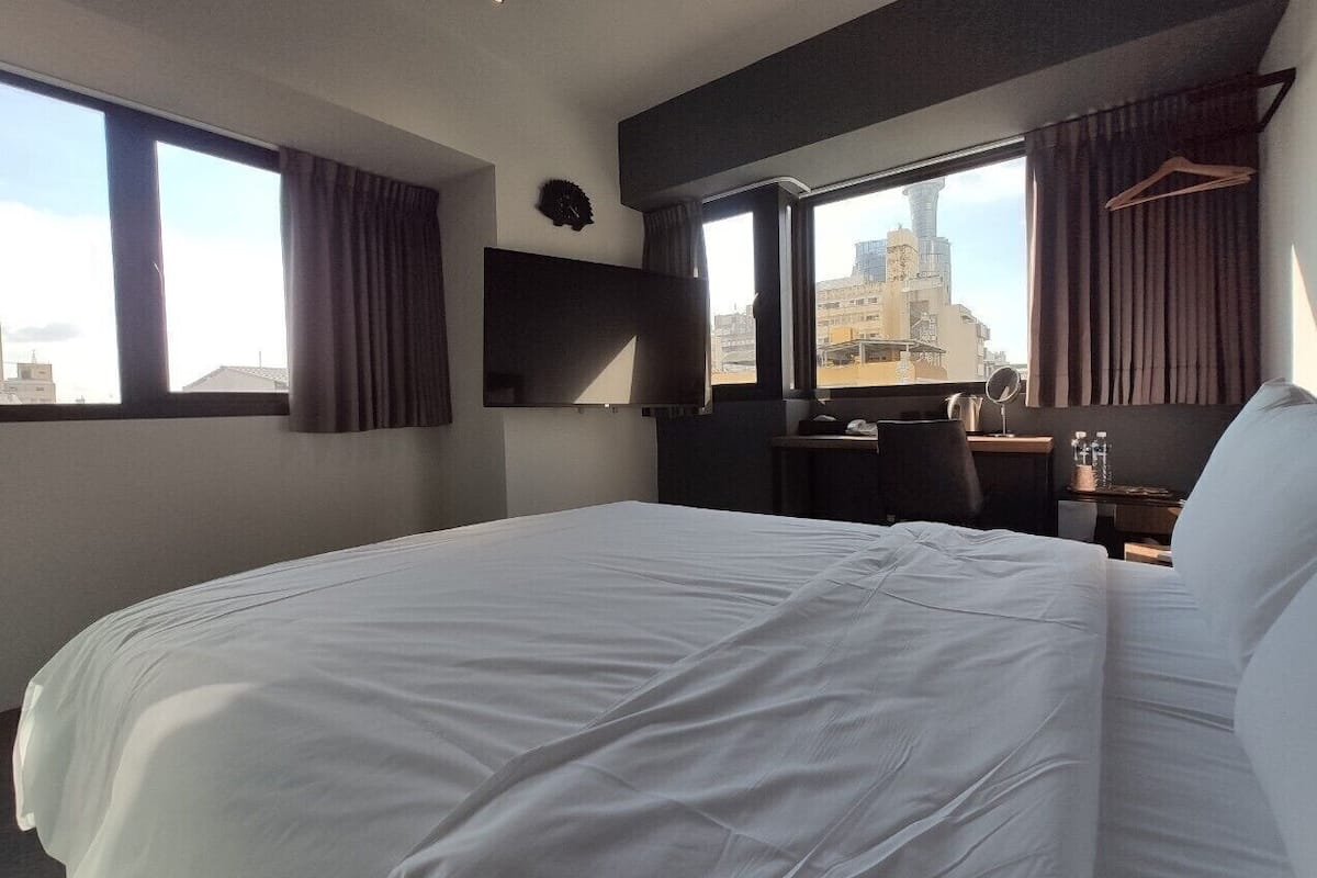 A hotel room with a large bed, a desk, a chair, a TV, and a view of the cityscape.