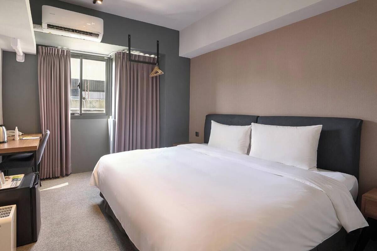 A hotel room with a large bed, a desk with a chair, an air conditioning unit, and a window with curtains.