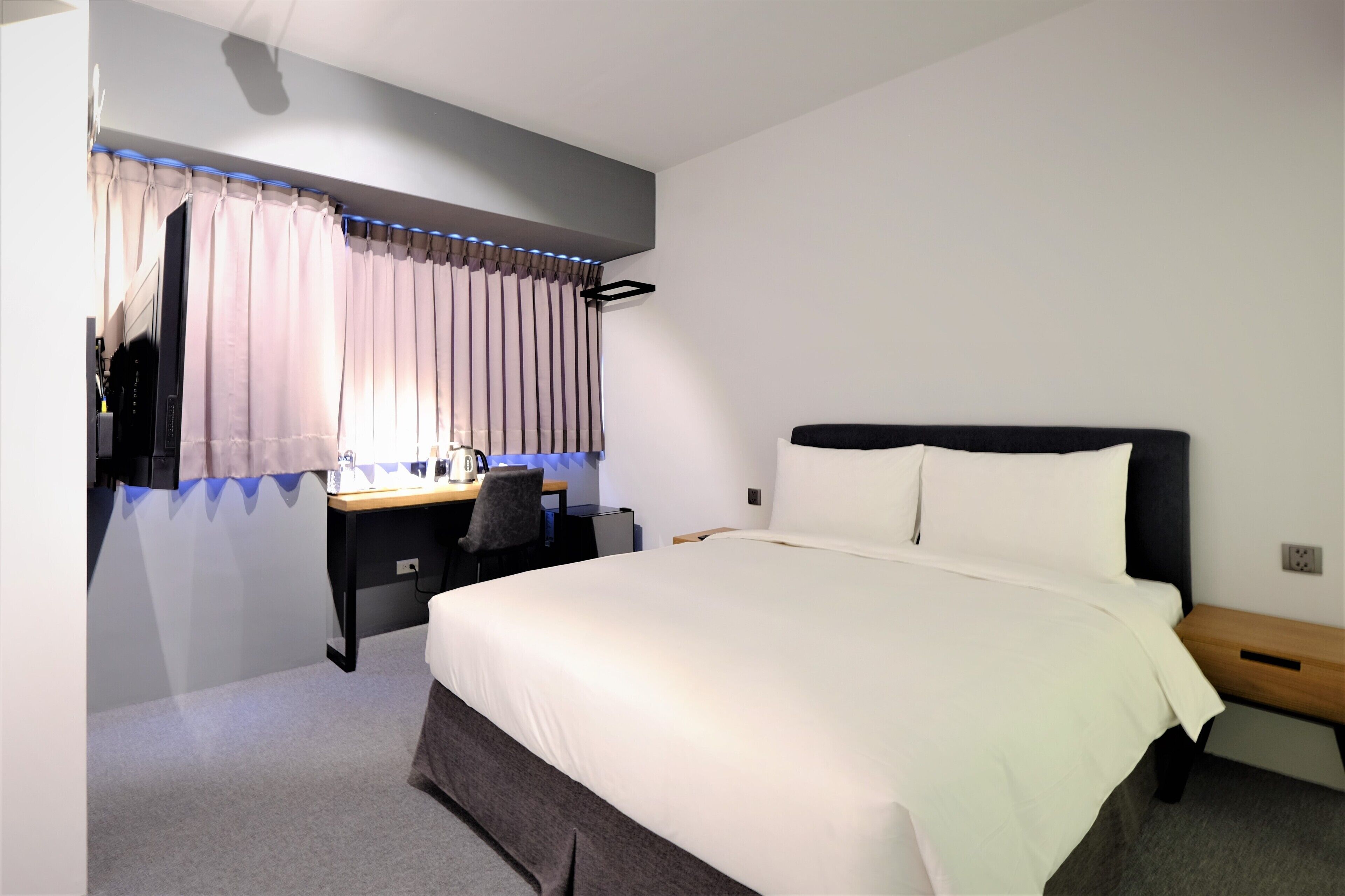 Ein Hotelzimmer mit einem großen Bett, einem Schreibtisch mit Stuhl, einem kleinen Tisch und einem Fenster mit Vorhängen.