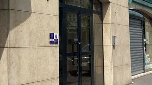 Rakennuksen julkisivussa on suuri lasinen sisäänkäyntiovi, sininen markiisi ja oikealla puolella suljettu sälekaihdin.